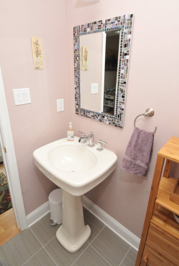 A small bathroom with a white pedestal sink, a mosaic-framed mirror, a lavender hand towel on a ring, soap dispenser, light pink walls, gray tile floor, and wooden shelving unit.