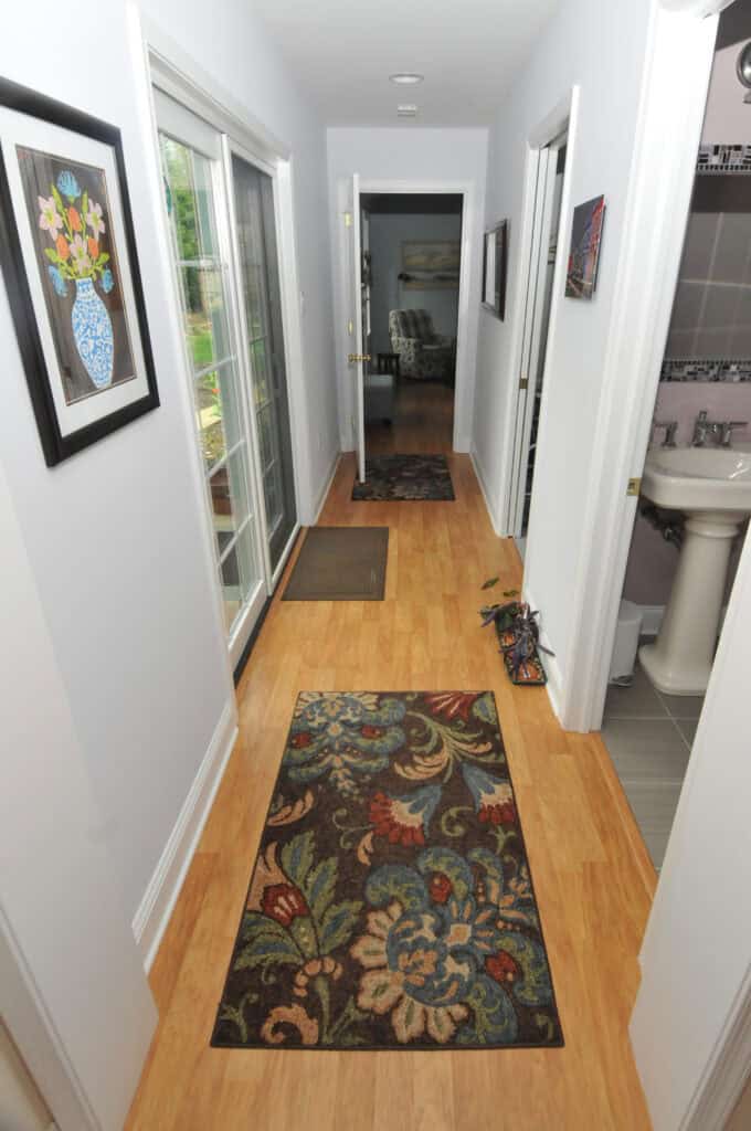 A hallway with light wood floors, two floral rugs, light-colored walls, framed artwork, and sliding glass doors on the left. The hallway leads to an open room, with a bathroom visible on the right.