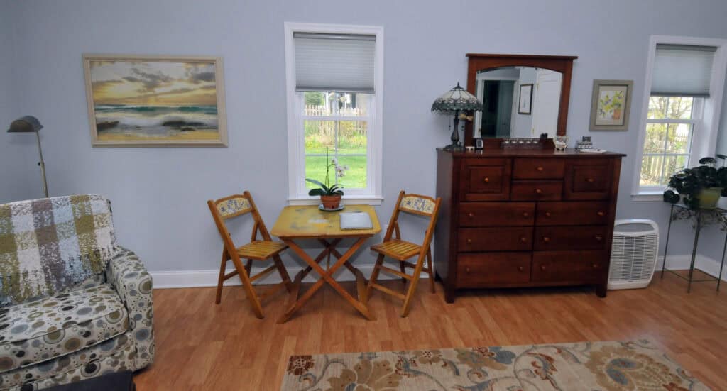 A cozy room with light blue walls, a small wooden table and two chairs, a dresser with a mirror, a floral armchair, a lamp, and framed art. Two windows let in natural light and show greenery outside.
