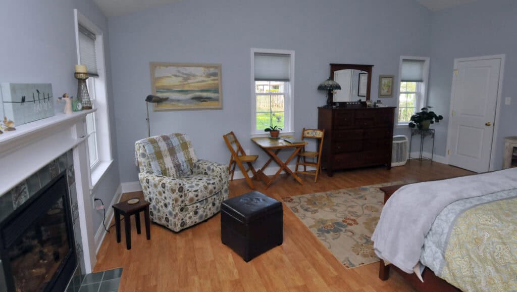 A cozy bedroom with light blue walls, a patterned armchair, a small table with two chairs, a wooden dresser with a mirror, a fireplace, and a bed with a gray comforter on a rug.