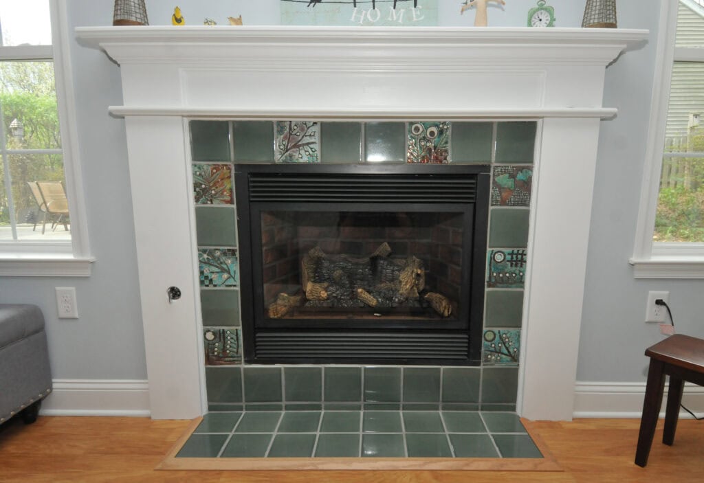 A fireplace with green decorative tiles, a black metal firebox, and a white wooden mantel, set against a light gray wall with windows and greenery visible outside.