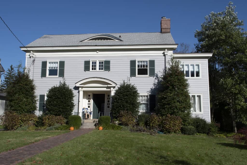 A two-story white house with green shutters and a gray roof sits behind a lawn. Two large bushes frame the front door, and two pumpkins are on the walkway. A person and a dog are on the porch. The sky is clear and blue.