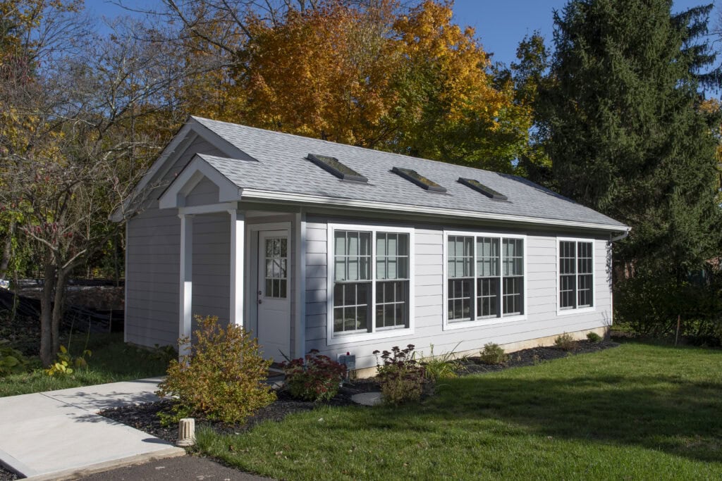A small, white cottage with large windows and skylights sits on a green lawn, surrounded by trees with autumn foliage under a clear blue sky.