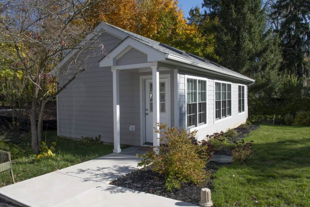 A small gray house with white trim, a front porch, and large windows sits among green grass and shrubs, with tall trees and autumn foliage in the background.