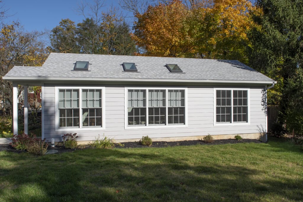 A white house with large windows and skylights stands in a green yard, surrounded by trees with autumn foliage under a clear blue sky.