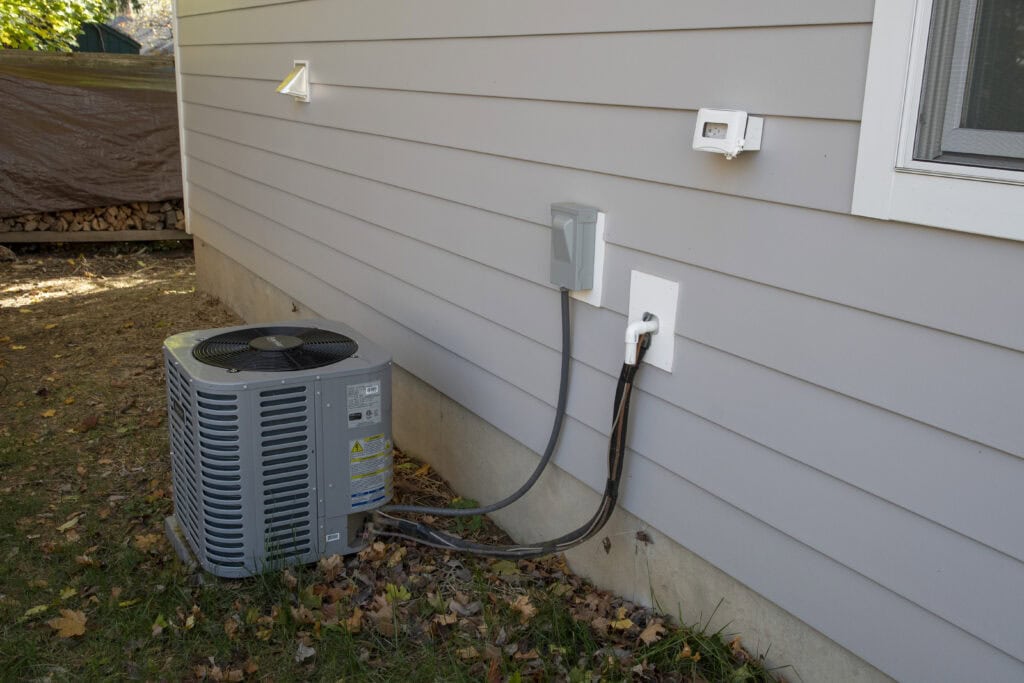 Outdoor air conditioning unit and electrical components mounted on the side of a gray house, with cables running into the wall and fallen leaves scattered on the ground.