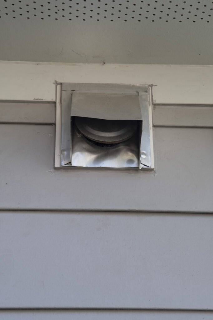 A close-up of a metal exterior vent cover attached to the side of a light gray house, under an overhang with perforated soffit panels.