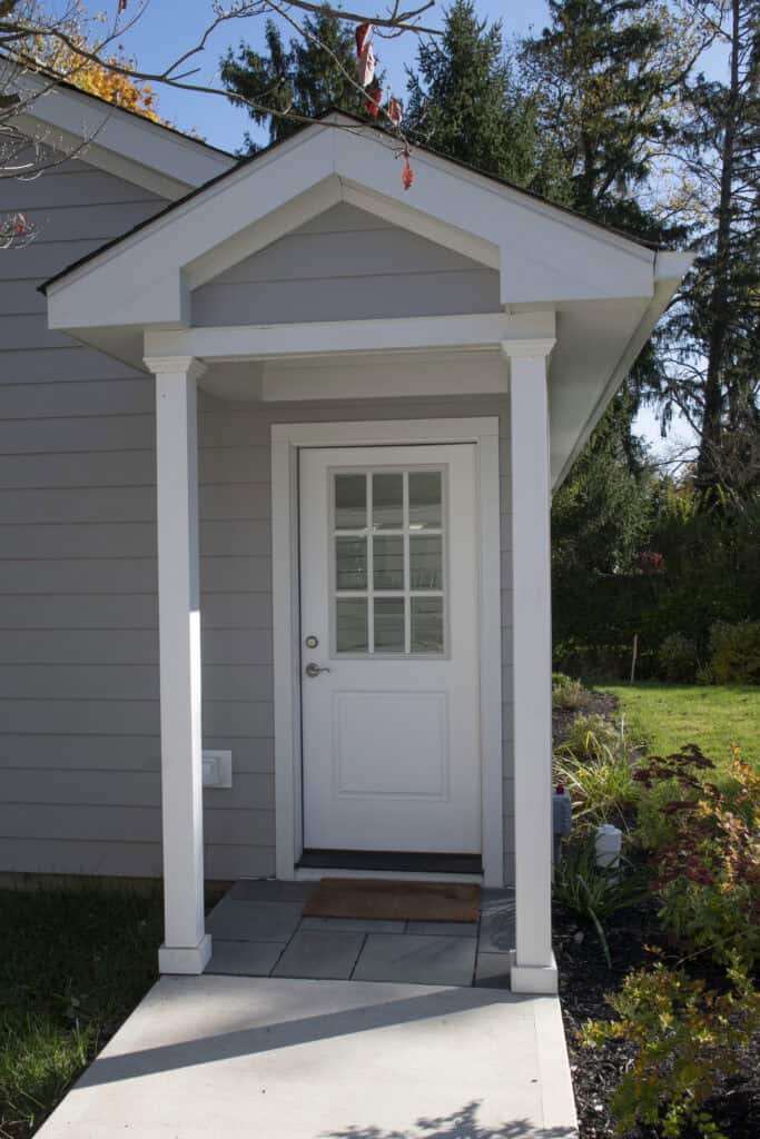 A white exterior door with glass panes is centered under a small porch with white columns and a triangular roof. The house has light gray siding, and landscaping with grass and shrubs surrounds the entryway.