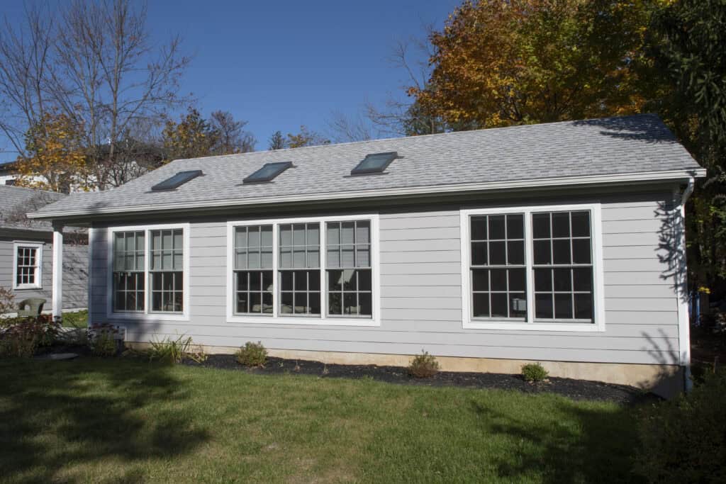 A single-story house with light gray siding, three large windows, and three skylights on the roof, sits on a green lawn with small shrubs, under a clear blue sky and surrounded by autumn trees.