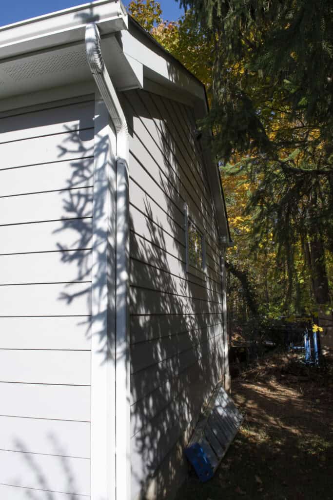 Side view of a light gray house with white trim and a gutter, partially shaded by tree branches. The sun casts leaf shadows on the siding, and there are building materials on the ground near the house.