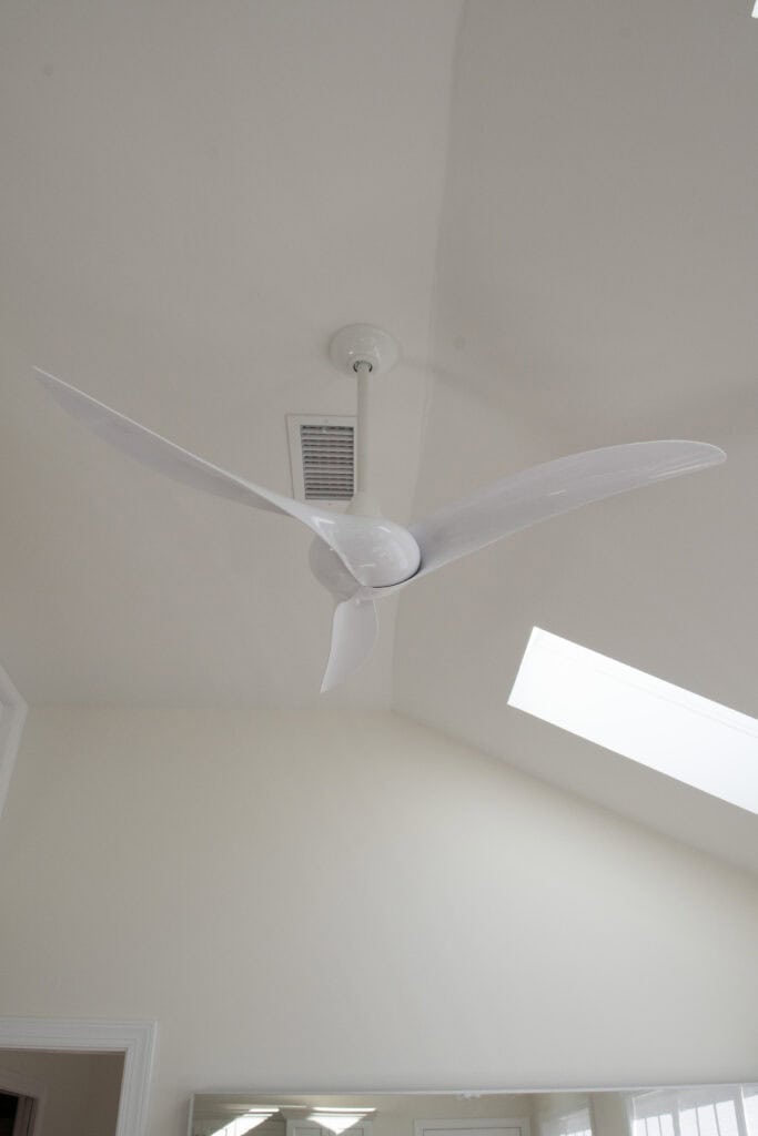 A modern white ceiling fan with three blades is mounted on a cream-colored ceiling. Nearby are a rectangular air vent and a skylight, letting in natural light.
