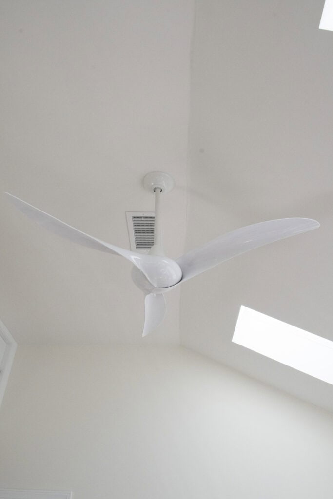 A white ceiling fan with three blades is mounted on a white ceiling. There is an air vent and two rectangular skylights nearby.