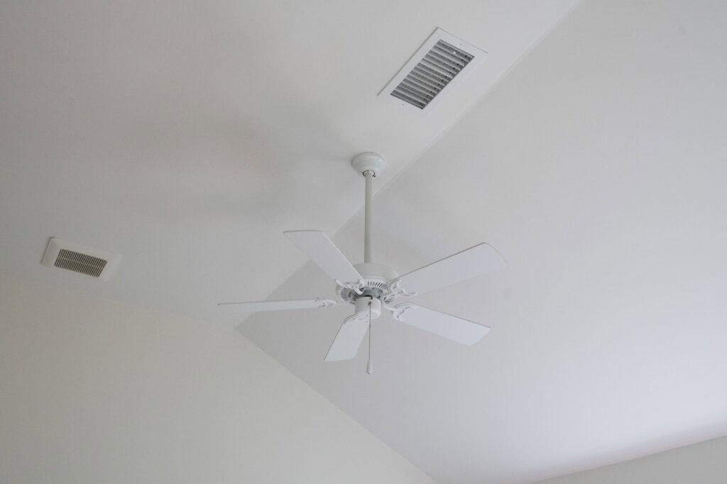 A white ceiling fan with five blades is mounted on a white ceiling. Two rectangular air vents are also visible nearby on the ceiling.