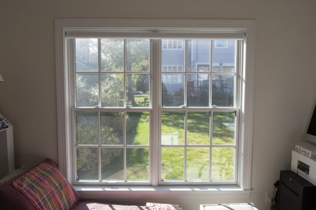 A large window with white framing lets sunlight into a room, showing a view of a green lawn, trees, and neighboring houses outside. Inside, part of a couch and furniture are visible near the window.