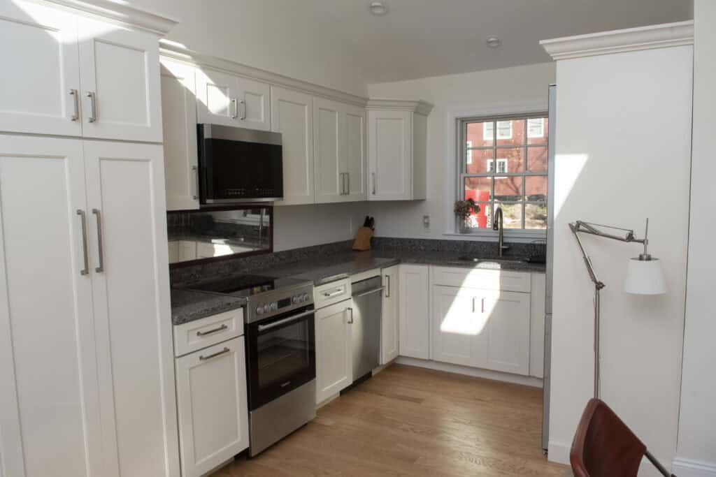 Modern kitchen with white cabinets, stainless steel appliances, a granite countertop, and a window letting in natural light. A lamp and chair are visible in the corner, and a knife block sits on the counter.