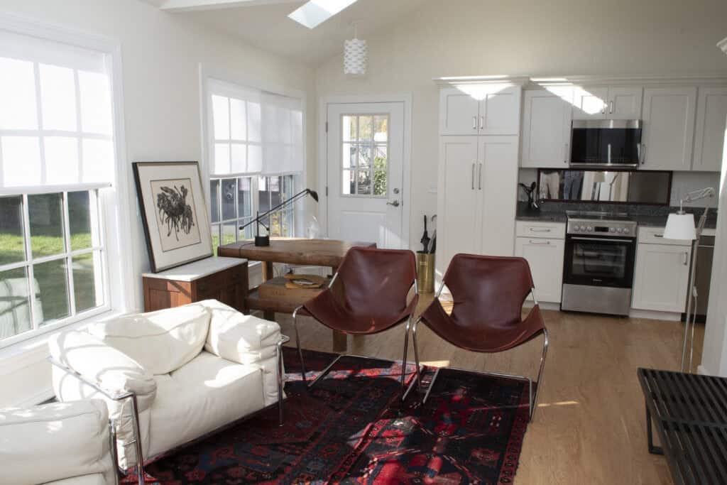 A bright, modern living room with large windows, a white armchair, two brown leather chairs, a red patterned rug, a wooden table, and a white kitchen area with appliances in the background.