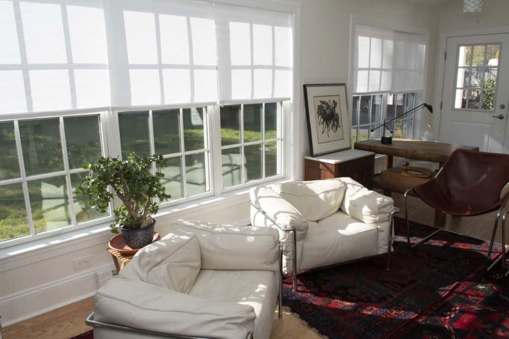 A sunlit room with large windows, two white armchairs, a brown leather chair, a wooden desk with a lamp, a potted plant, and framed artwork on the wall over a red patterned rug.