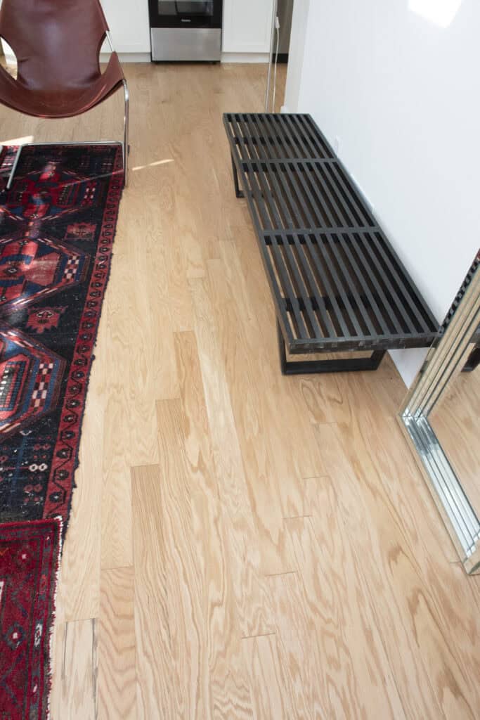 A wooden floor with a patterned red rug, a brown chair, a black slatted bench, and a large mirror leaning against the white wall in a modern room.