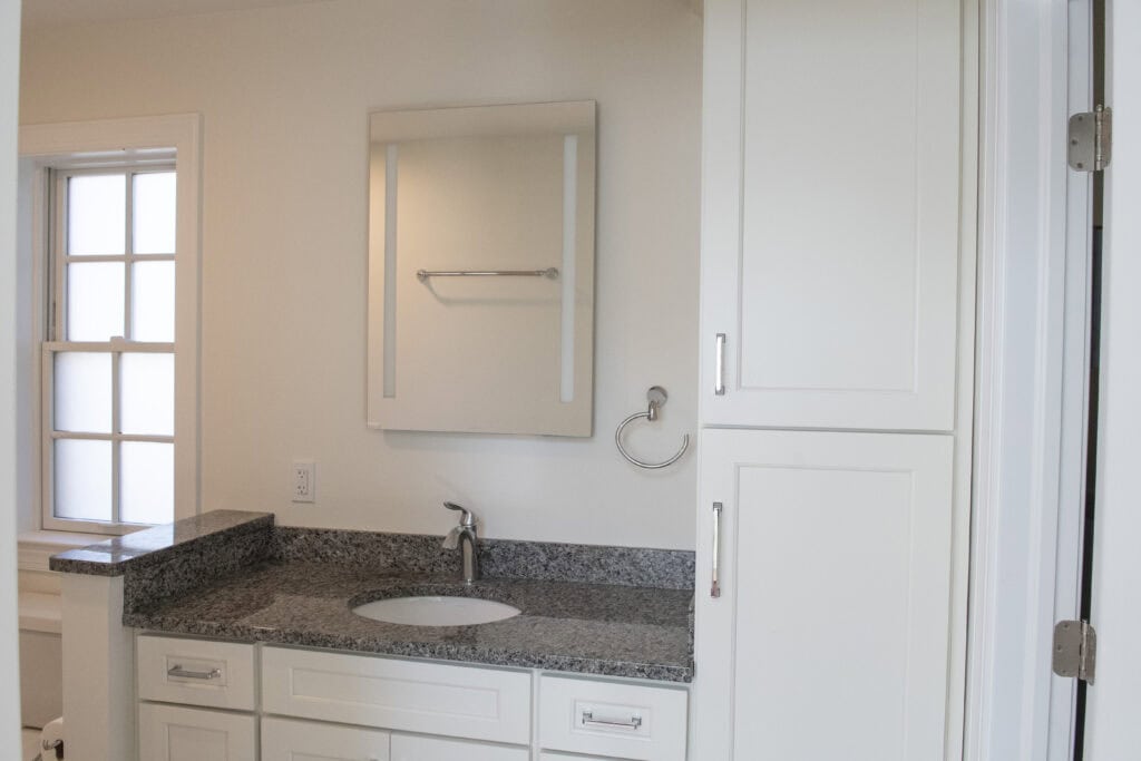 A clean, modern bathroom with white cabinets, a granite countertop with a sink, a wall-mounted mirror, and a window with frosted glass panes.