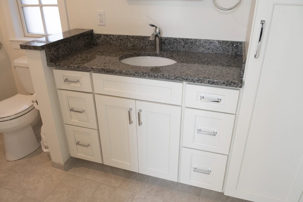 White bathroom vanity with granite countertop, an under-mount sink, chrome faucet, and silver handles. The vanity is next to a toilet, tiled floor, and white walls with natural light from a window.