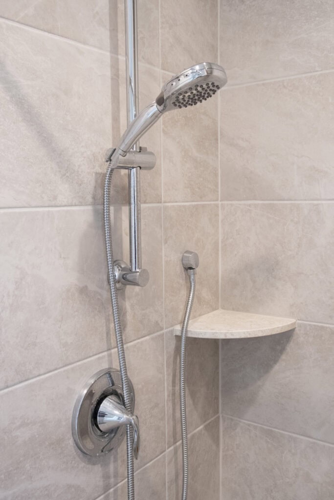 A modern shower with a metal handheld showerhead, adjustable wall-mounted holder, and a flexible hose, installed on beige tiled walls with a small corner shelf.