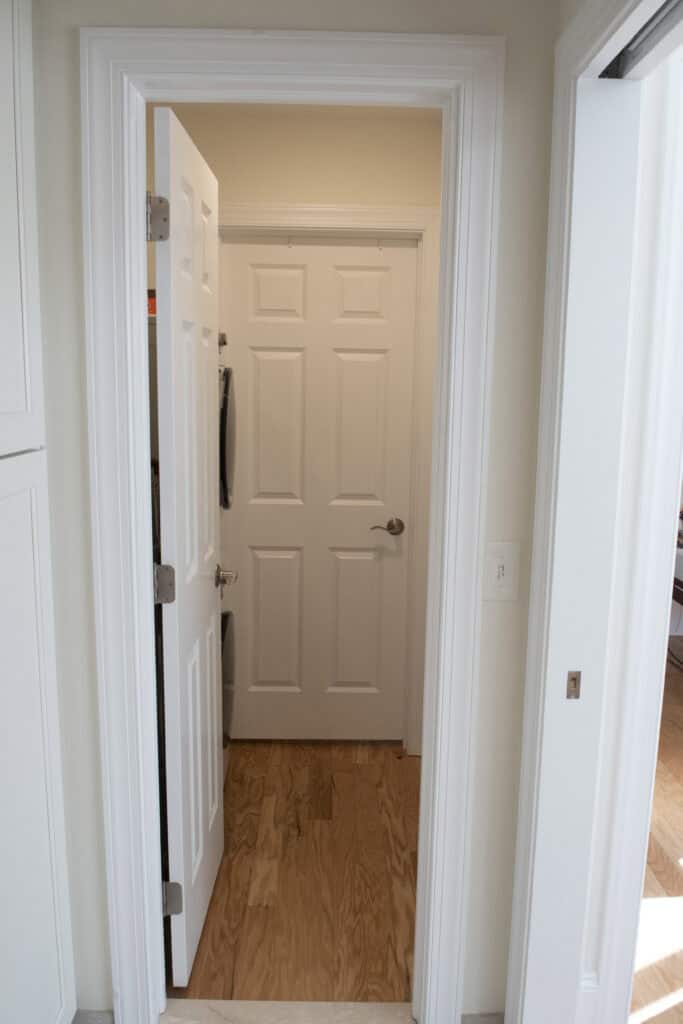 A white door is partially open, revealing another closed white door in a hallway with light-colored walls and wood flooring.