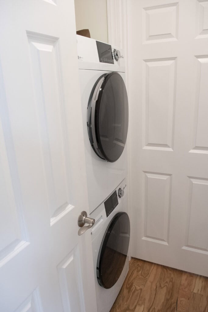 A stacked washer and dryer unit is placed in a small laundry closet with white folding doors and a wooden floor.