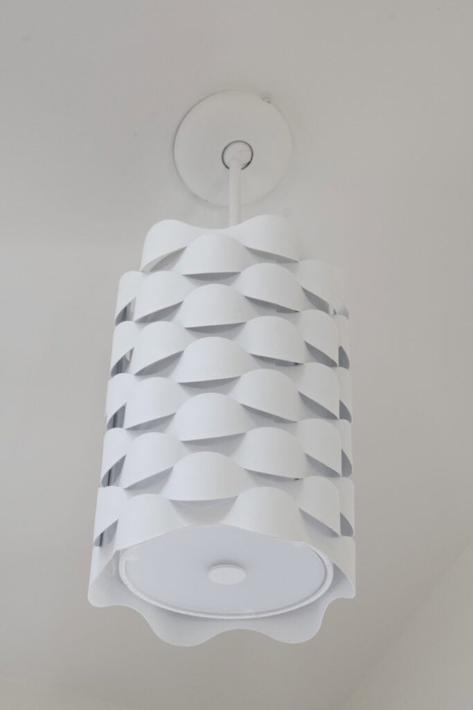 A modern pendant light fixture with a cylindrical, wavy white shade featuring a layered, overlapping pattern, hanging from a white ceiling.