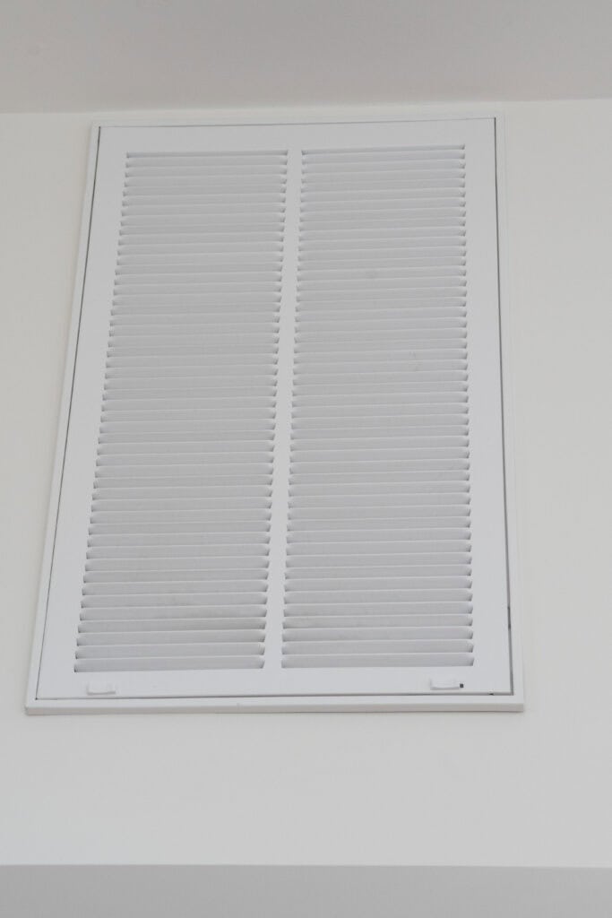 A large white air vent or return grille is mounted on a plain white wall, with horizontal slats covering the vent opening.