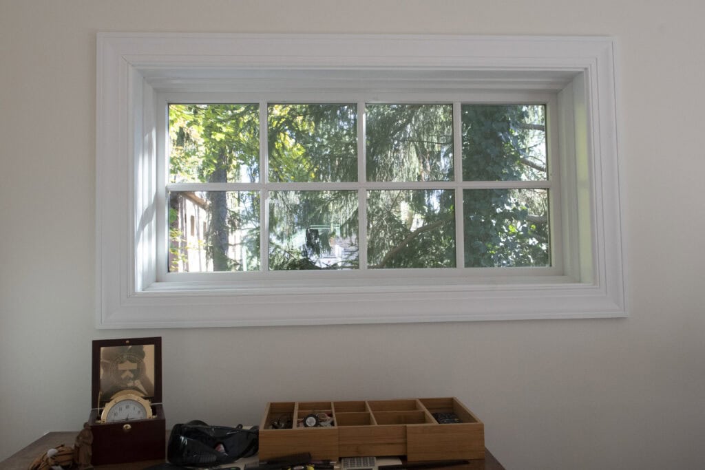 A white-framed window shows green trees outside. Below the window, a wooden desk holds a wooden organizer with small items, a clock, and various personal belongings.