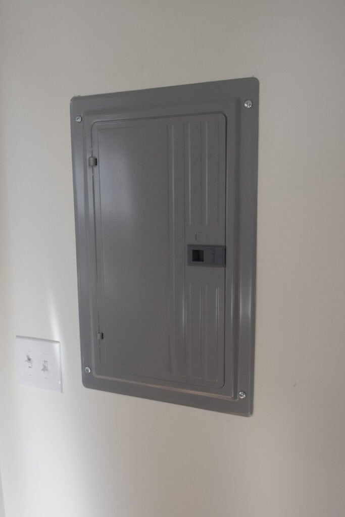 A closed gray electrical breaker panel is mounted on a white wall, with a light switch panel featuring two switches located to the left of the breaker box.