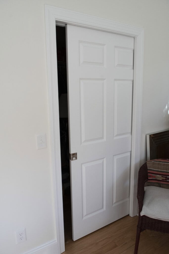 A white sliding closet door is partially open in a room with light-colored walls and a wooden floor. A brown chair with a cushion and a framed picture are next to the door.