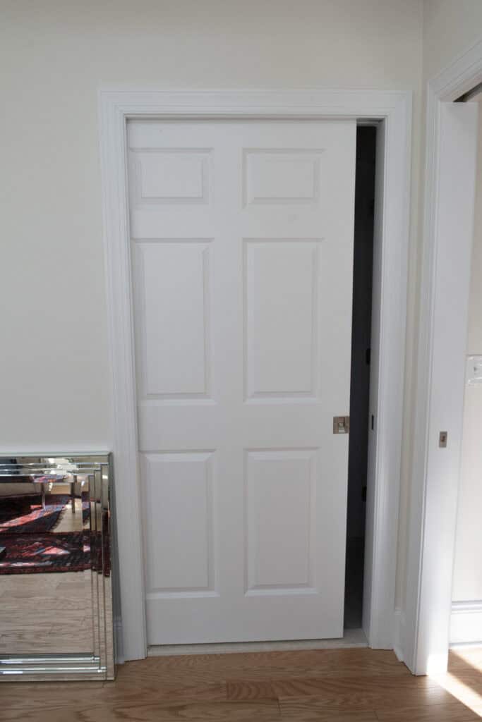 A white sliding pocket door is partially open in a room with light-colored walls and a wooden floor. A mirrored cabinet and part of a colorful rug are visible to the left of the door.