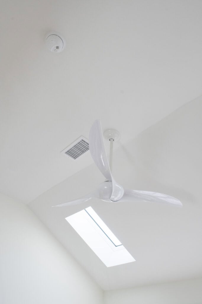 A modern white ceiling fan with three blades is mounted on a white ceiling, near a smoke detector, an air vent, and a rectangular skylight that lets in natural light.