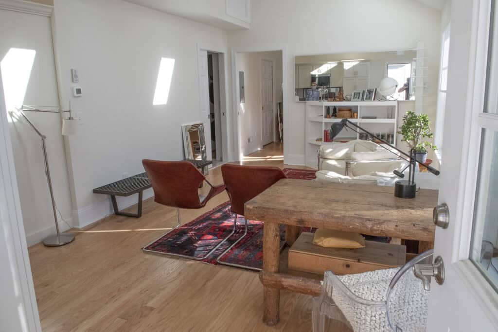 Bright, modern room with wooden floors, featuring a rustic desk, two brown chairs on a red rug, a bench, shelves with decor, a floor lamp, and a large mirror reflecting the space.