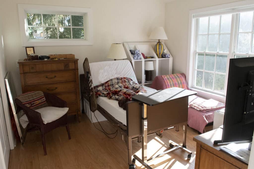 A sunlit room with a hospital bed covered by a plaid blanket, a wooden dresser, armchair, side table with a lamp, pink armchair by a window, and a TV on a stand. The floor is wood and the room looks tidy.