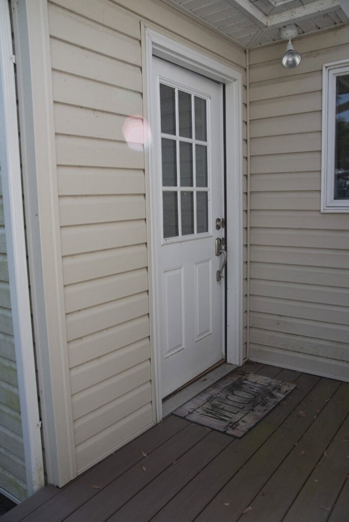 A white door with glass panels is set in beige siding. A “Welcome” doormat is in front of the door on a wooden porch. A small outdoor light fixture hangs above to the right of the door.