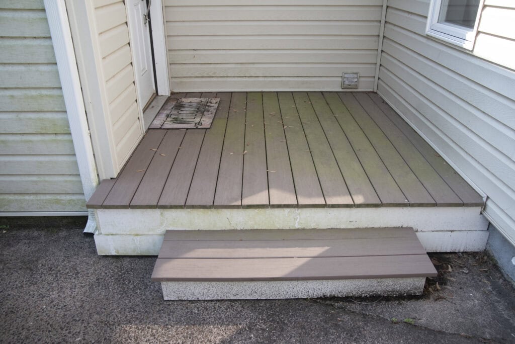 A small wooden porch with a single step, beige siding, a door mat by the door, and some scattered debris on the deck. Part of the porch shows green mildew or algae stains.
