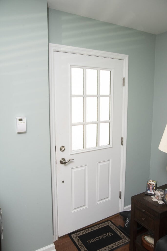 A white door with six frosted glass panels is set in a light teal wall. There is a thermostat on the wall, a small table with a lamp and photos, and a doormat that says Pawsome upside down.