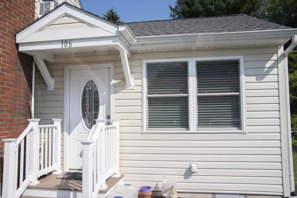 Small white house entrance with oval glass door, covered porch, white railings, steps, two large windows with closed blinds, and brick wall on the left. House number 106 is displayed above the door.