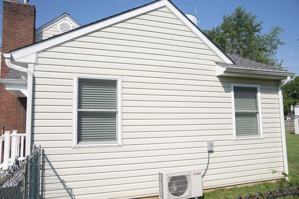 A beige house with horizontal vinyl siding, two windows with closed blinds, and an outdoor air conditioning unit mounted at the base. There is a brick chimney on the left and a grassy yard on the right.