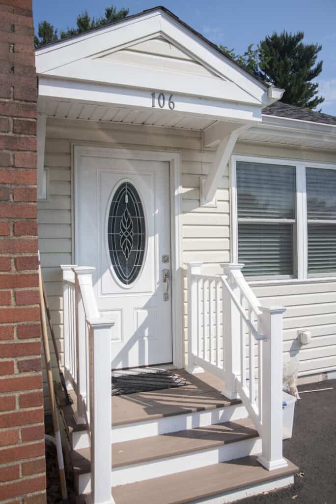 A small front porch with white railings and steps leads to a white door with an oval glass panel on a beige house. The house number 106 is above the door. A doormat sits at the entrance.