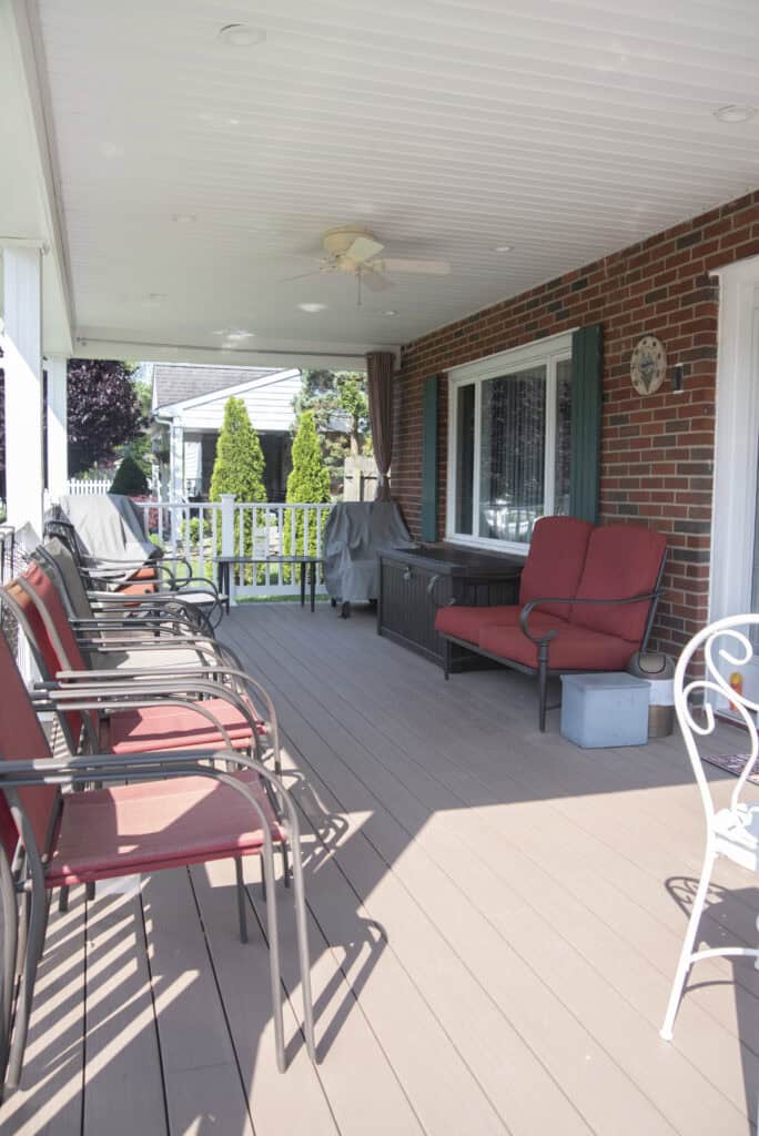 A covered porch with red cushioned chairs lined along a wall, a ceiling fan overhead, and a grill in the corner. The space overlooks a green garden and white fence.