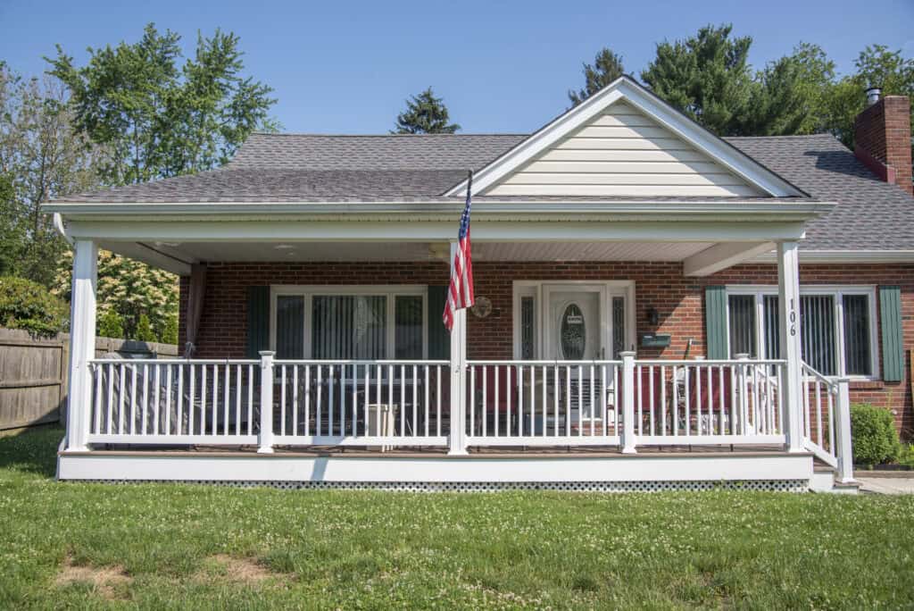 A single-story brick house with a covered front porch, white railing, green shutters, and an American flag hanging near the entrance. The yard is grassy with a wooden fence and trees in the background.