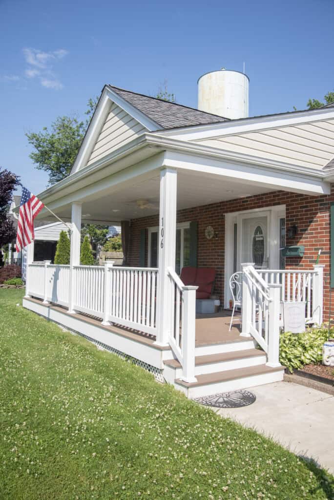 A brick house with a covered front porch, white railing, and an American flag. Patio chairs and a small table are on the porch. The house number 106 is visible, and there is green grass in the yard under a clear blue sky.