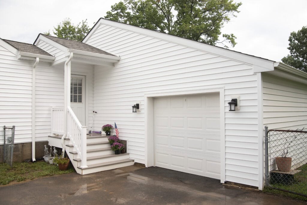A white house with a single-car garage, two black outdoor lights, a small porch with steps, potted plants, and an American flag decoration near the door. A chain-link fence borders the driveway.