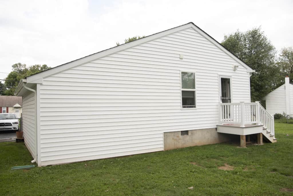 A small white house with vinyl siding, a concrete foundation, a single window, and a back door with a small porch and steps, set on a grassy lawn. Trees and another house are visible in the background.