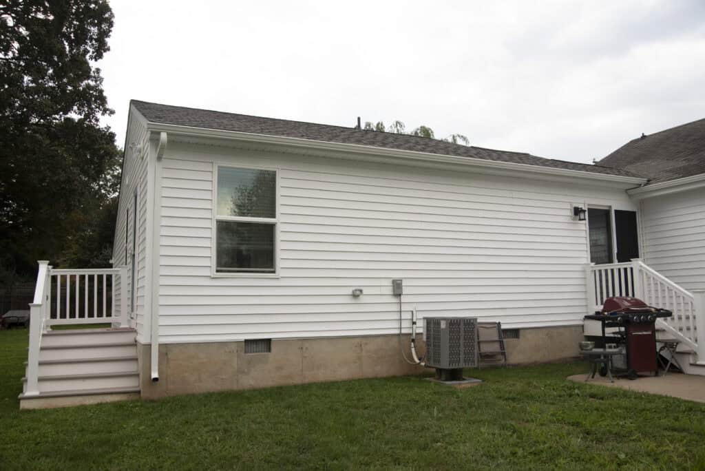 A white single-story house with vinyl siding, a small porch with stairs, a window, an outdoor air conditioning unit, and a red grill on a concrete patio in the backyard. The grass lawn is green and well-kept.
