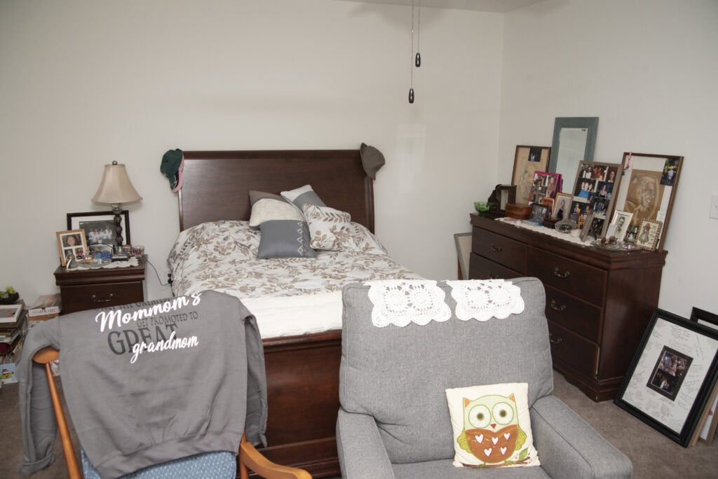 A cozy bedroom with a made bed, two gray armchairs in front, a dresser with family photos, and a framed picture on the floor. A sweatshirt reading Mormons favorite grandmom hangs on one chair.