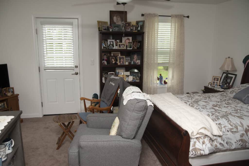 A cozy bedroom with a gray armchair, a wooden rocking chair, and a neatly made bed. A bookshelf full of framed photos stands by two windows with sheer curtains, and a small table sits beside the chairs.